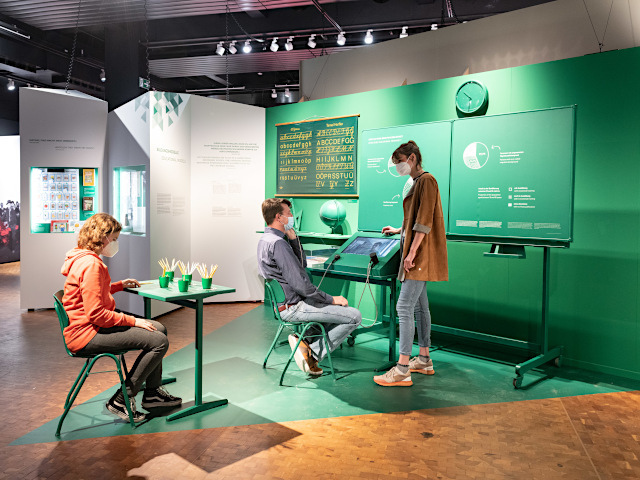 Besucher sitzen in einem nachgebauten Klassenzimmer in grüner Farbe
